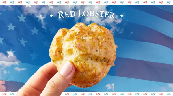 A hand holding a cheddar bay biscuit over a background of clouds and the United States flag. An eagle is superimposed on the biscuit